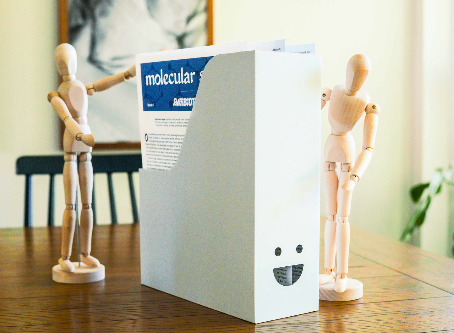 White cardboard box with a face-shaped cutout on the front, which holds several paper publications. It sists on a table with two wooden mannequins gathered around it.