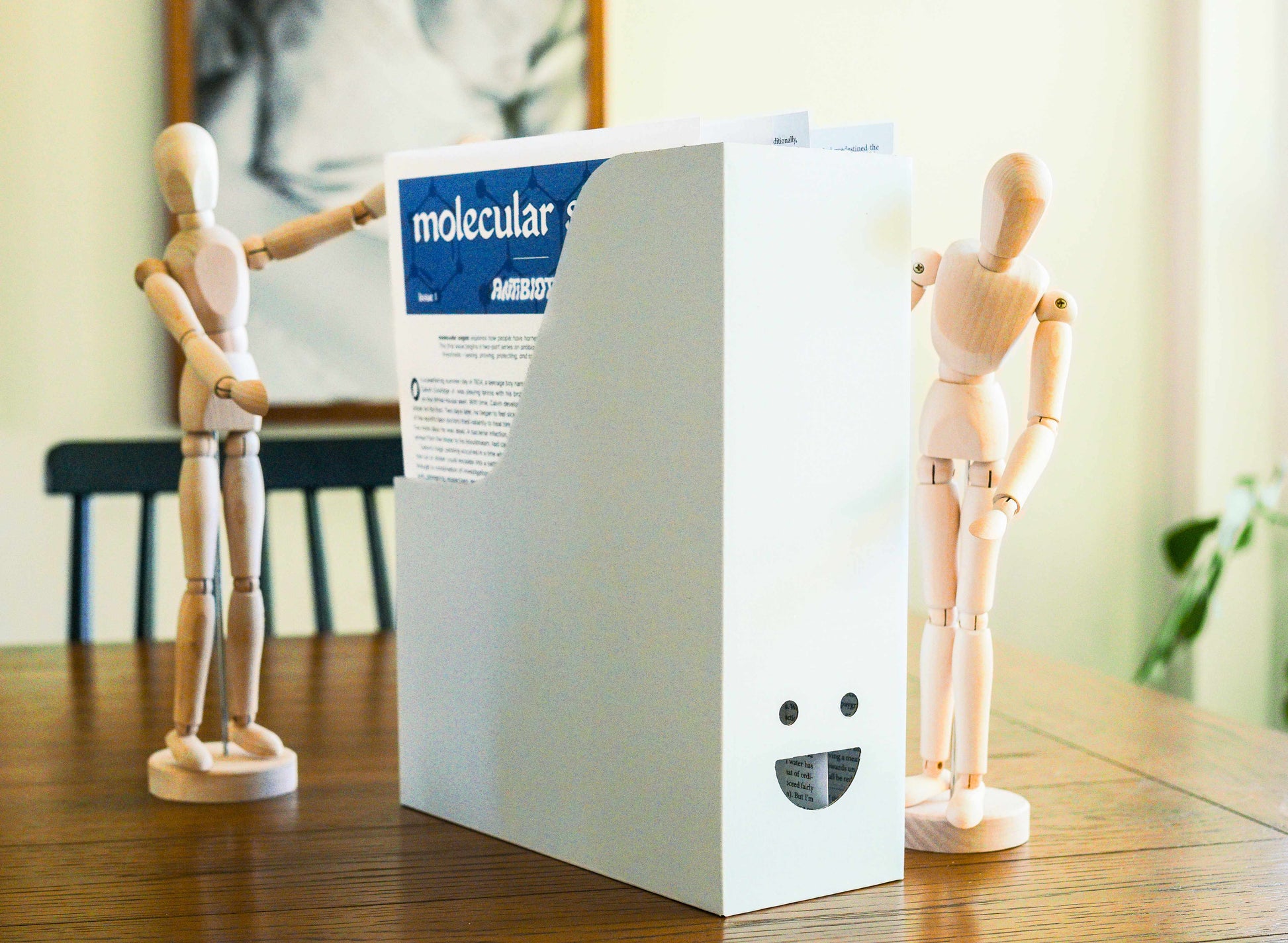 White cardboard box with a face-shaped cutout on the front, which holds several paper publications. It sists on a table with two wooden mannequins gathered around it.