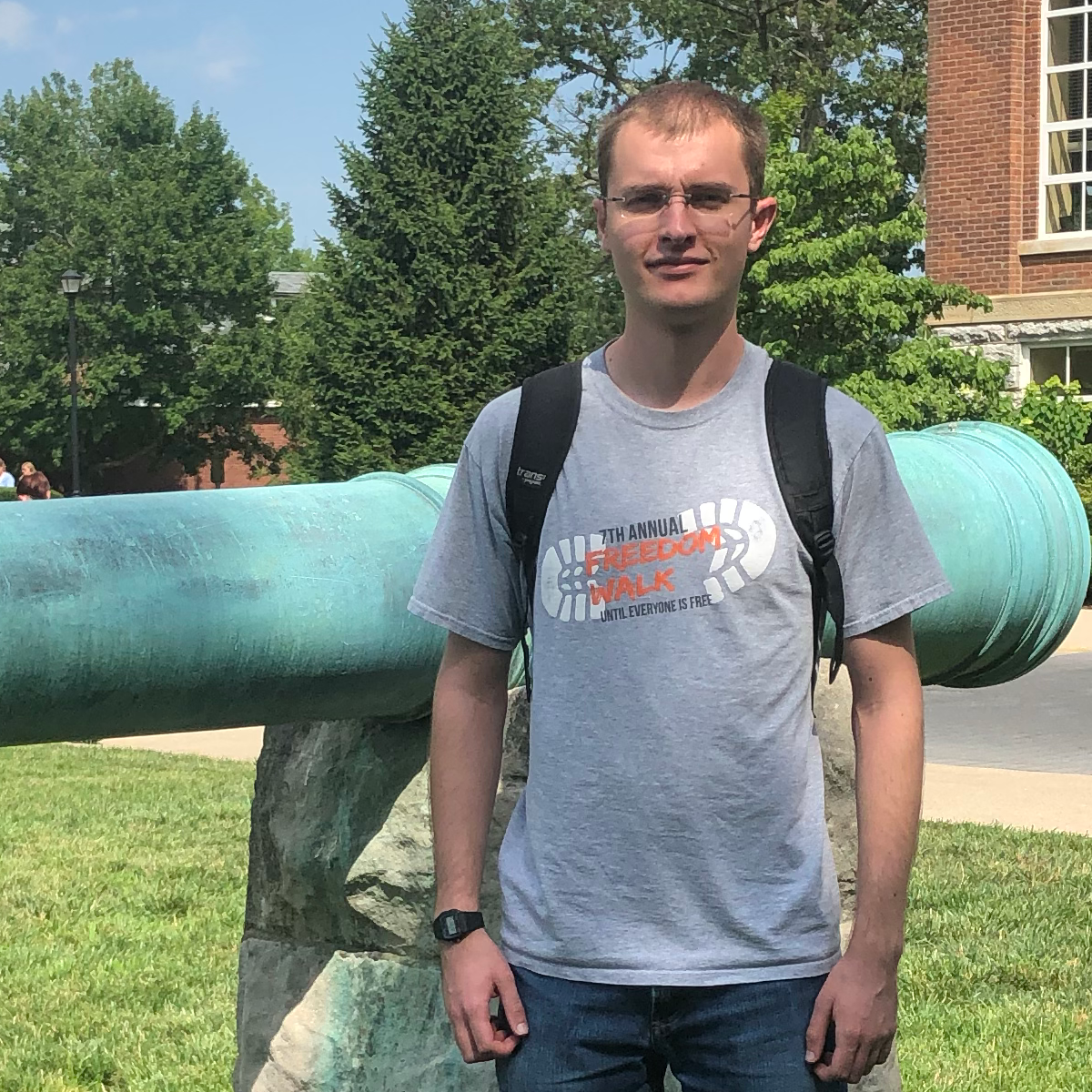 Man wearing a gray t-shirt standing in front of a green cannon barrel on a stone mount