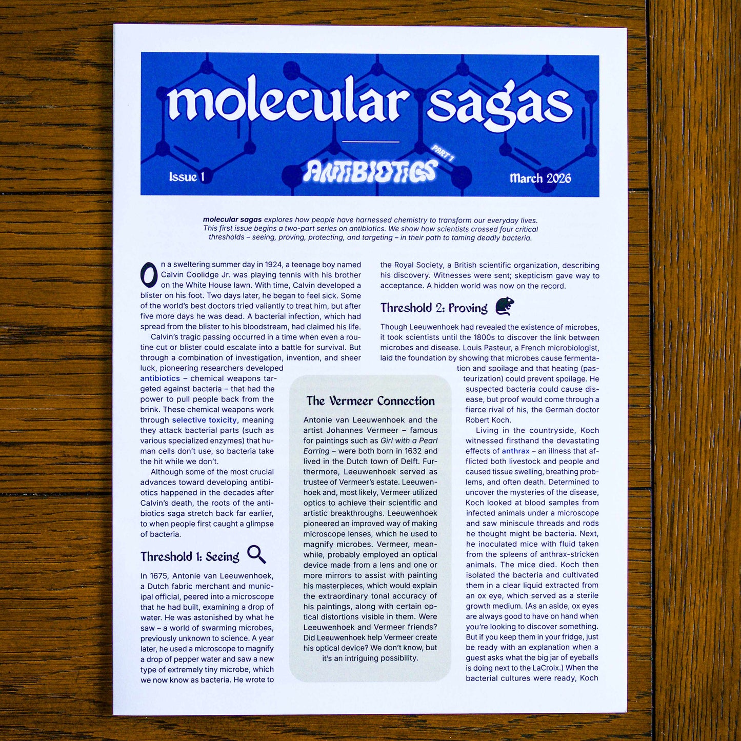 Paper publication titled 'molecular sagas' on a wooden background. Headings on the publication read 'Issue 1', 'Antibiotics Part 1', and 'March 2026'.