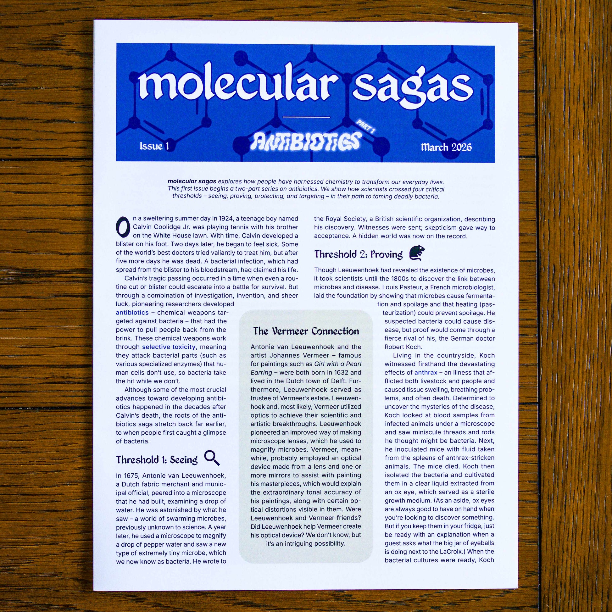 Paper publication titled 'molecular sagas' on a wooden background. Headings on the publication read 'Issue 1', 'Antibiotics Part 1', and 'March 2026'.