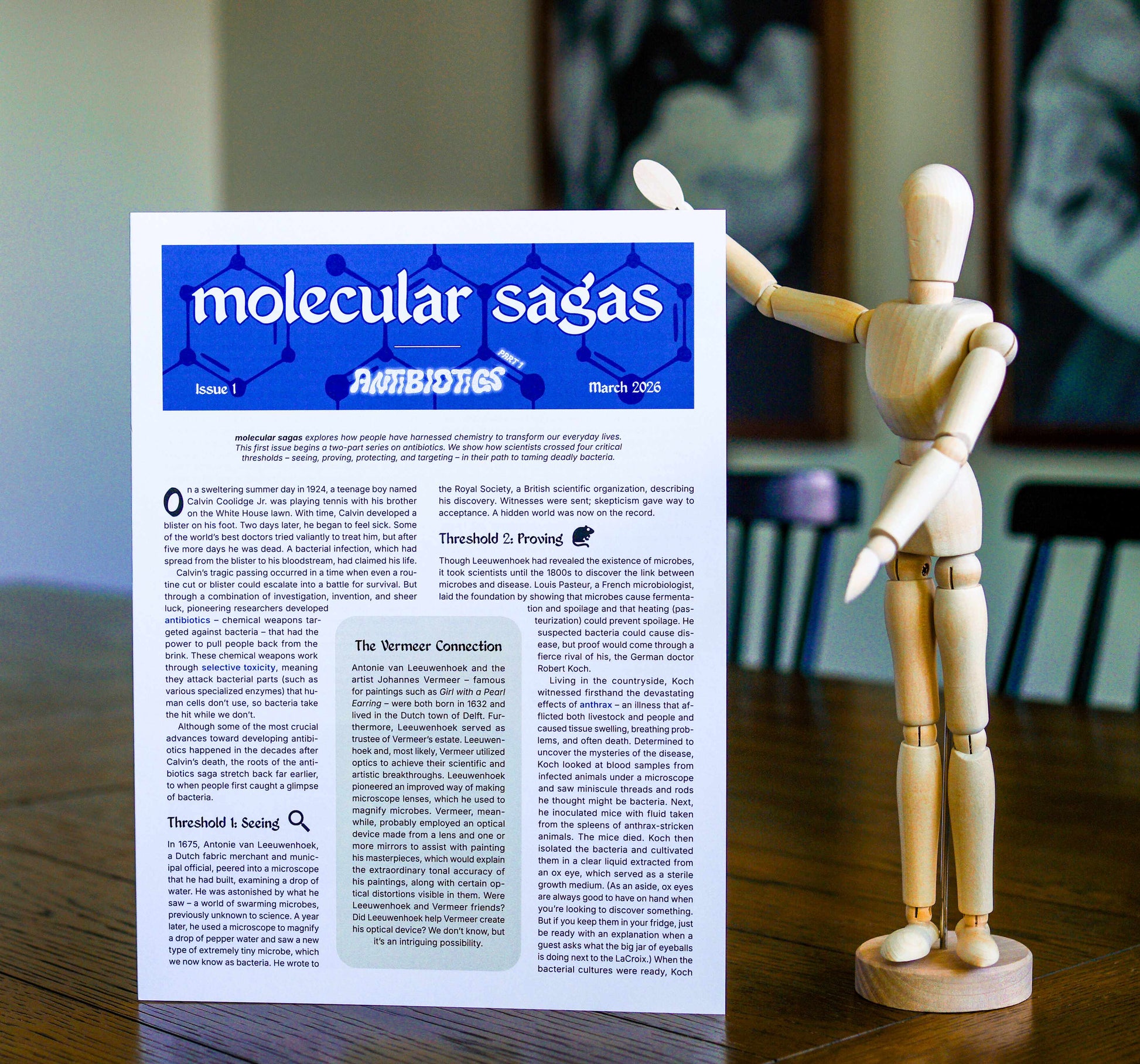 Paper publication titled 'molecular sagas' standing on a table with a wooden mannequin posing beside it. Headings on the publication read 'Issue 1', 'Antibiotics Part 1', and 'March 2026'.