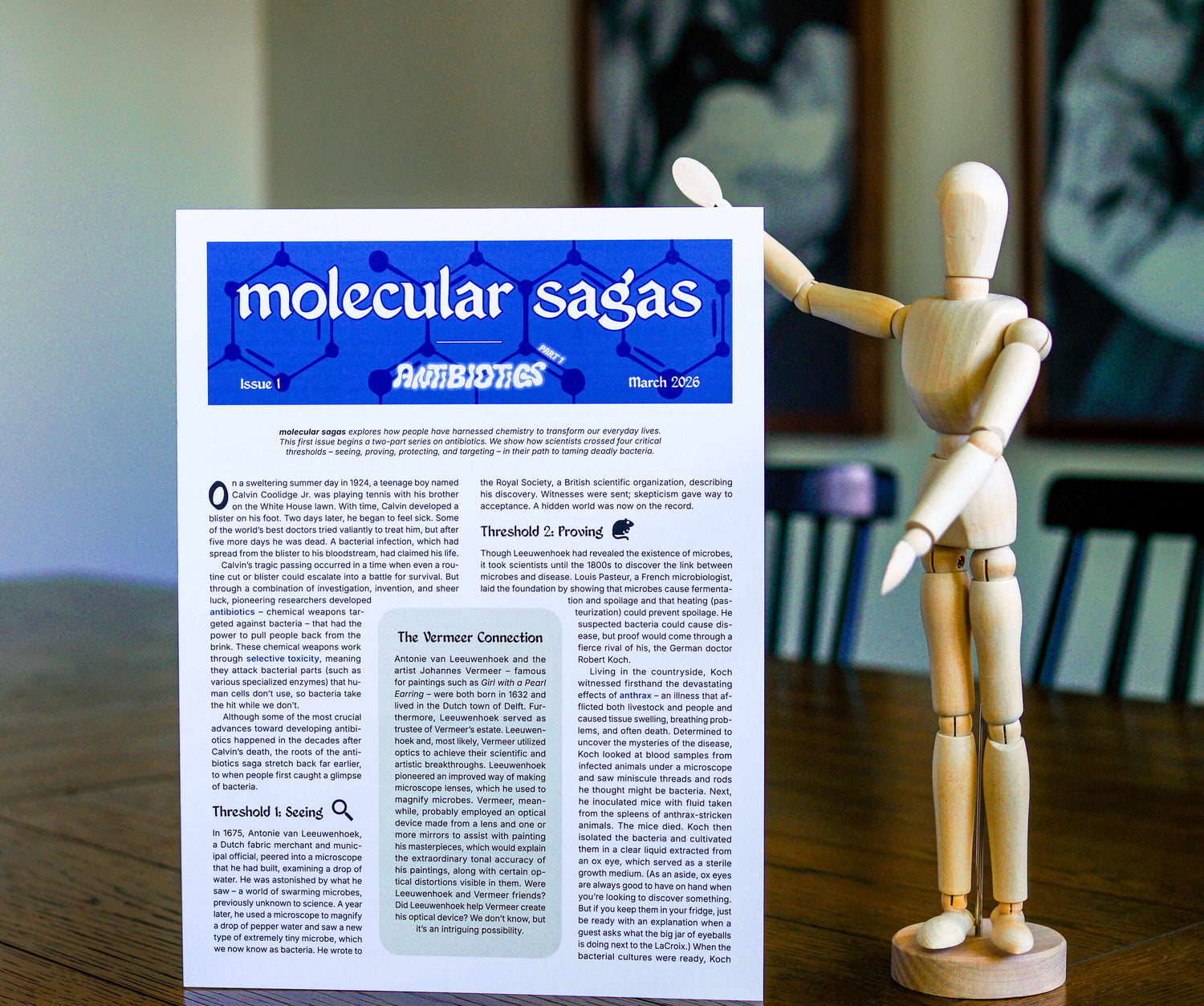 Paper publication titled 'molecular sagas' standing on a table with a wooden mannequin posing beside it. Headings on the publication read 'Issue 1', 'Antibiotics Part 1', and 'March 2026'.